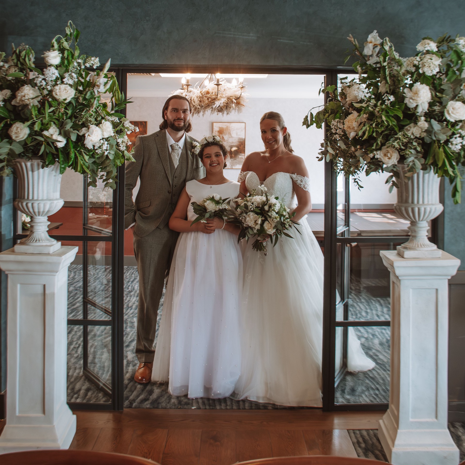 A bride, groom and young bridesmaid posing together indoors between two large floral arrangements