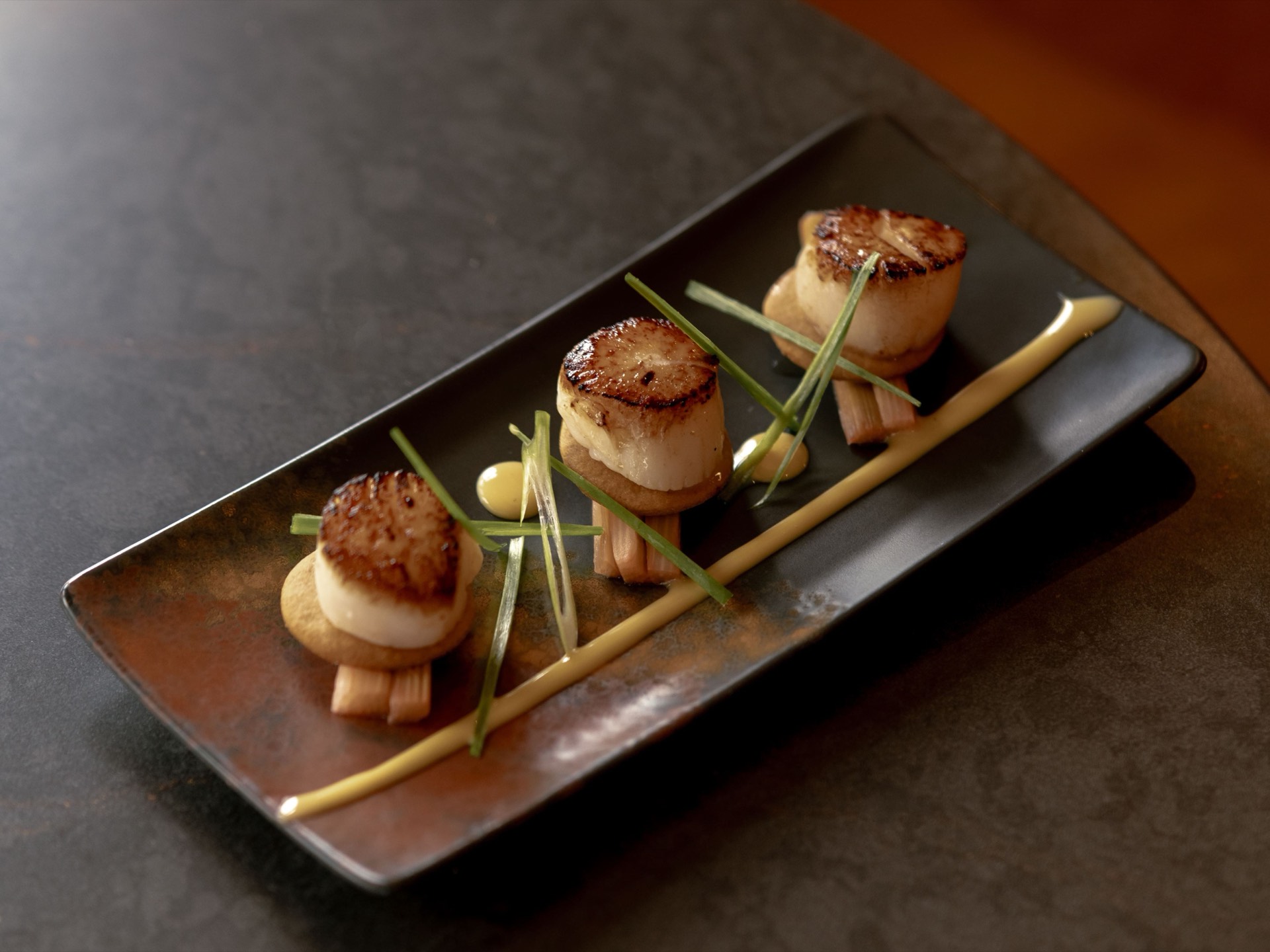 An elegant plate of seared scallops garnished with greens and sauce on a black rectangular dish