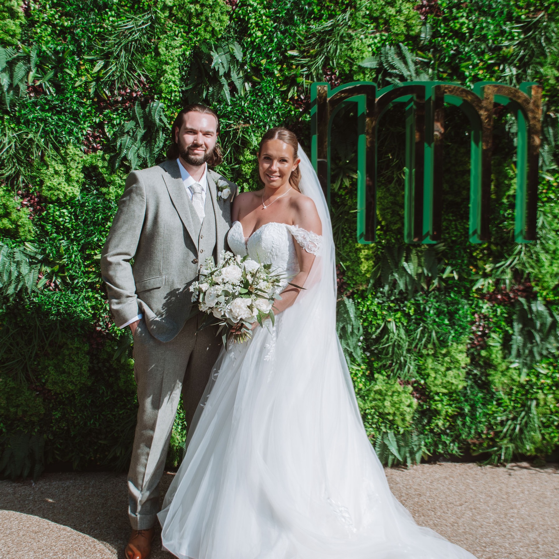 A bride and groom posing together outdoors in front of a lush green wall, the bride holding a bouquet