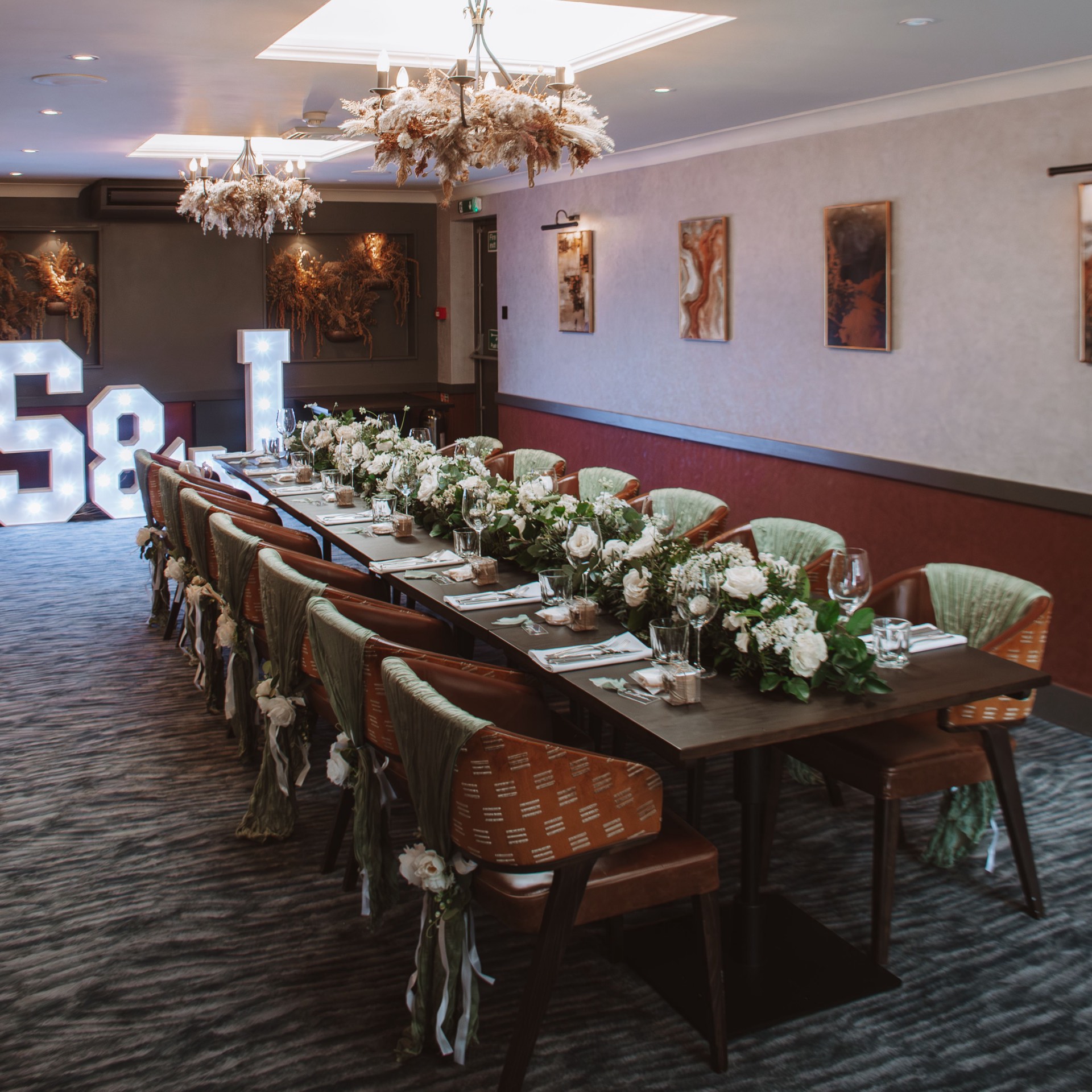 A long table decorated with white flowers, green fabric on the chairs and illuminated letters in the background